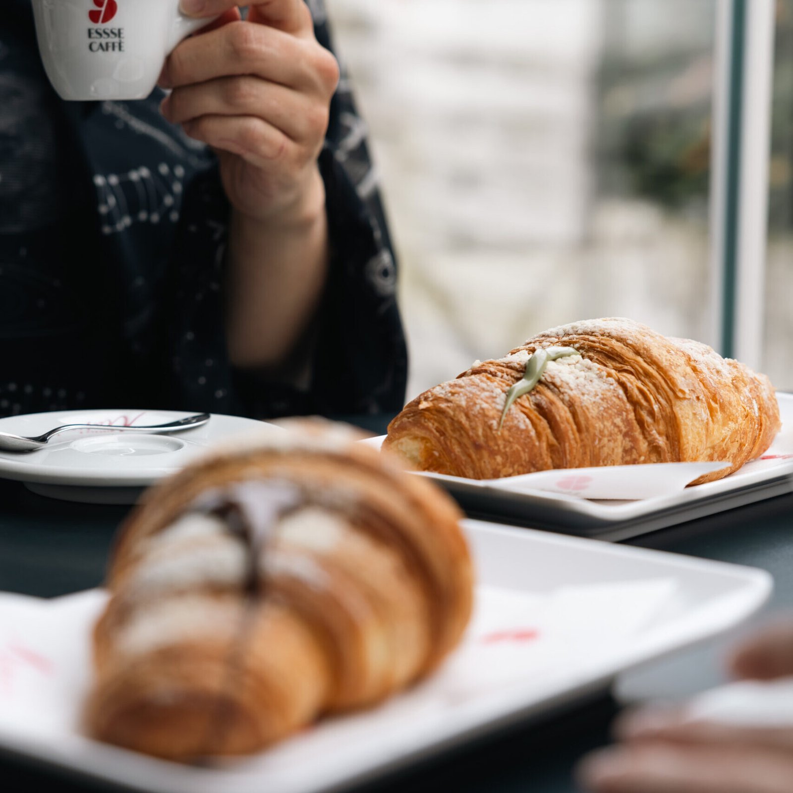 Maffia 1957 Una persona con in mano una tazza di caffè bianco siede a un tavolo con due croissant su piatti bianchi, uno dei quali condito con zucchero a velo e una guarnizione, godendo della morbida luce naturale vicino alla finestra di un'accogliente pasticceria di Como. Panetteria Pasticceria Caffetteria Como e Provincia