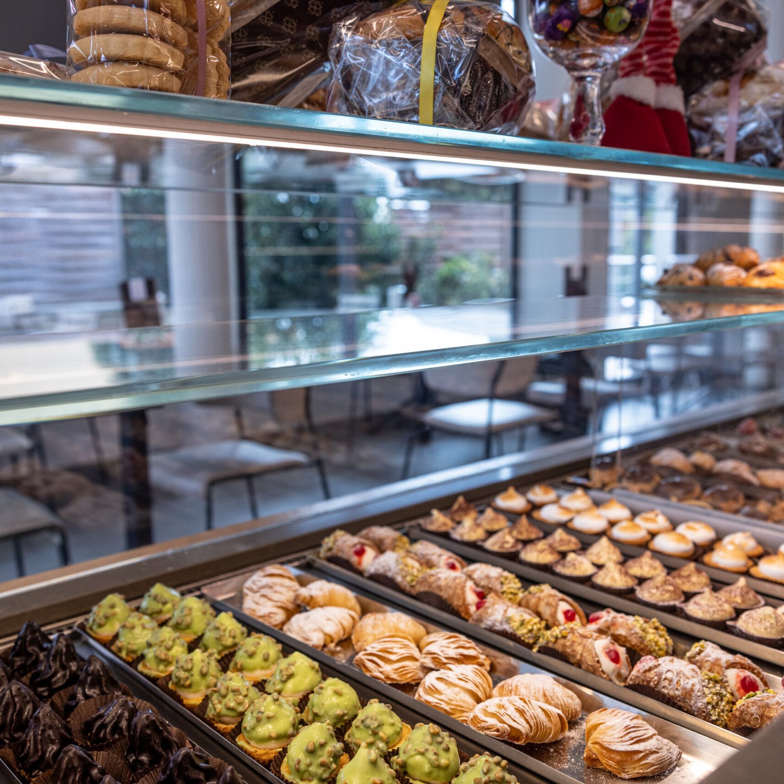Maffia 1957 Una vetrina in una pasticceria di Como mostra dolci assortiti, biscotti e dessert disposti in file. I ripiani sovrastanti contengono sacchetti di biscotti, mentre sullo sfondo sono visibili tavoli e sedie. Panetteria Pasticceria Caffetteria Como e Provincia