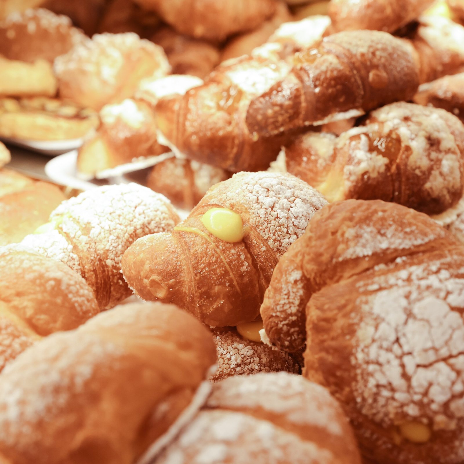 Maffia 1957 Primo piano di diversi croissant dorati spolverati di zucchero a velo e ripieni di crema pasticcera gialla, disposti in pila in una panetteria di Como, con altri dolci visibili sullo sfondo. Panetteria Pasticceria Caffetteria Como e Provincia