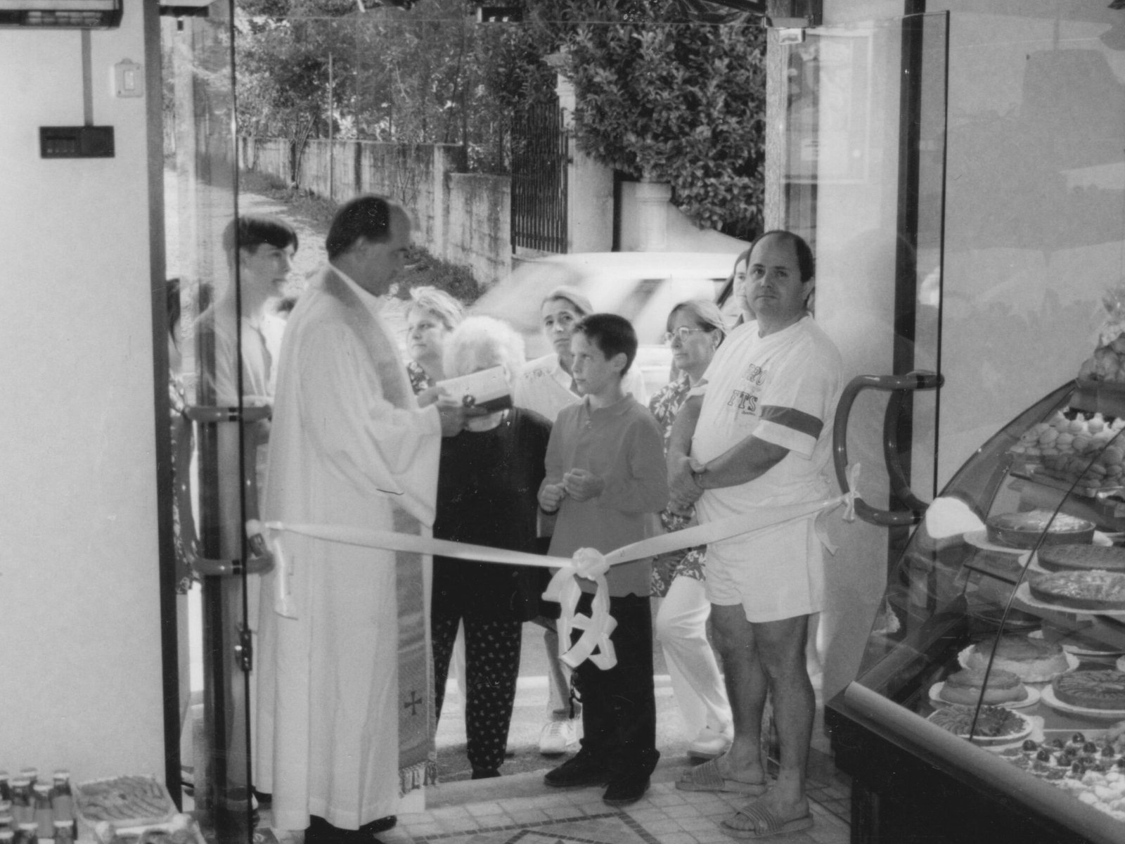 Maffia 1957 Un sacerdote si trova all'ingresso di una panetteria artigianale di Como e benedice un gruppo riunito per il taglio del nastro. Pasticcini e torte sono visibili in una vetrina sulla destra. Panetteria Pasticceria Caffetteria Como e Provincia