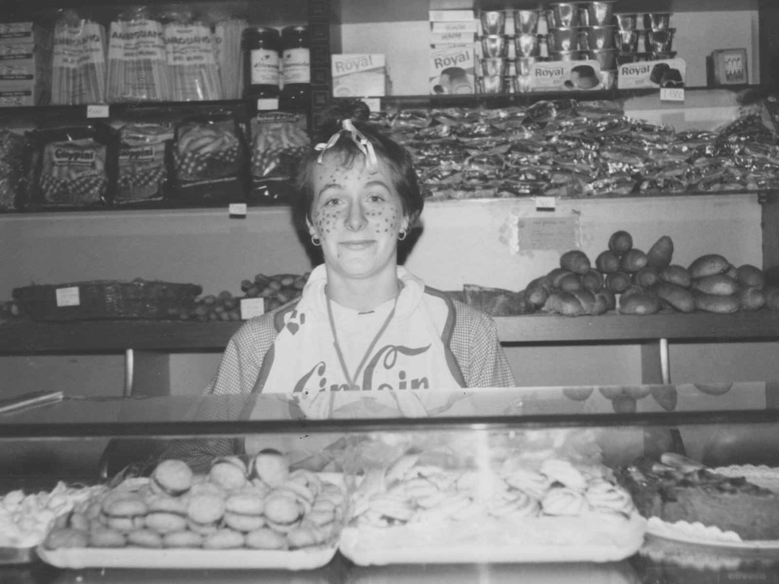 Maffia 1957 Una persona con un foulard in testa e le lentiggini dipinte sul viso sta dietro il bancone della panetteria di Como, circondata da delizioso pane artigianale comasco e scaffali di leccornie, pronta a servire i clienti. Panetteria Pasticceria Caffetteria Como e Provincia