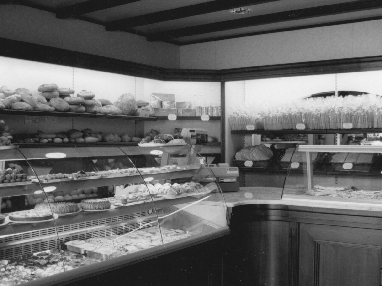 Maffia 1957 Foto in bianco e nero di un interno di un panificio como con scaffali e vetrine pieni di vari pani, dolci e prodotti da forno. Piccoli cartellini dei prezzi sono visibili sugli scaffali e sui banconi. Panetteria Pasticceria Caffetteria Como e Provincia