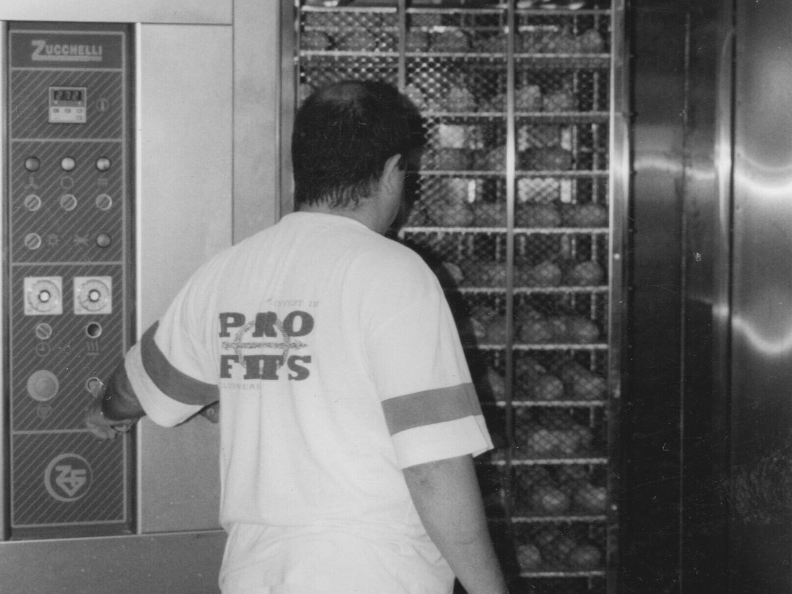 Maffia 1957 Una persona in maglietta e pantaloncini sta davanti a un grande forno industriale di un panificio di Como, premendo un pulsante sul pannello di controllo, con vassoi di cibo visibili all'interno del forno. Panetteria Pasticceria Caffetteria Como e Provincia