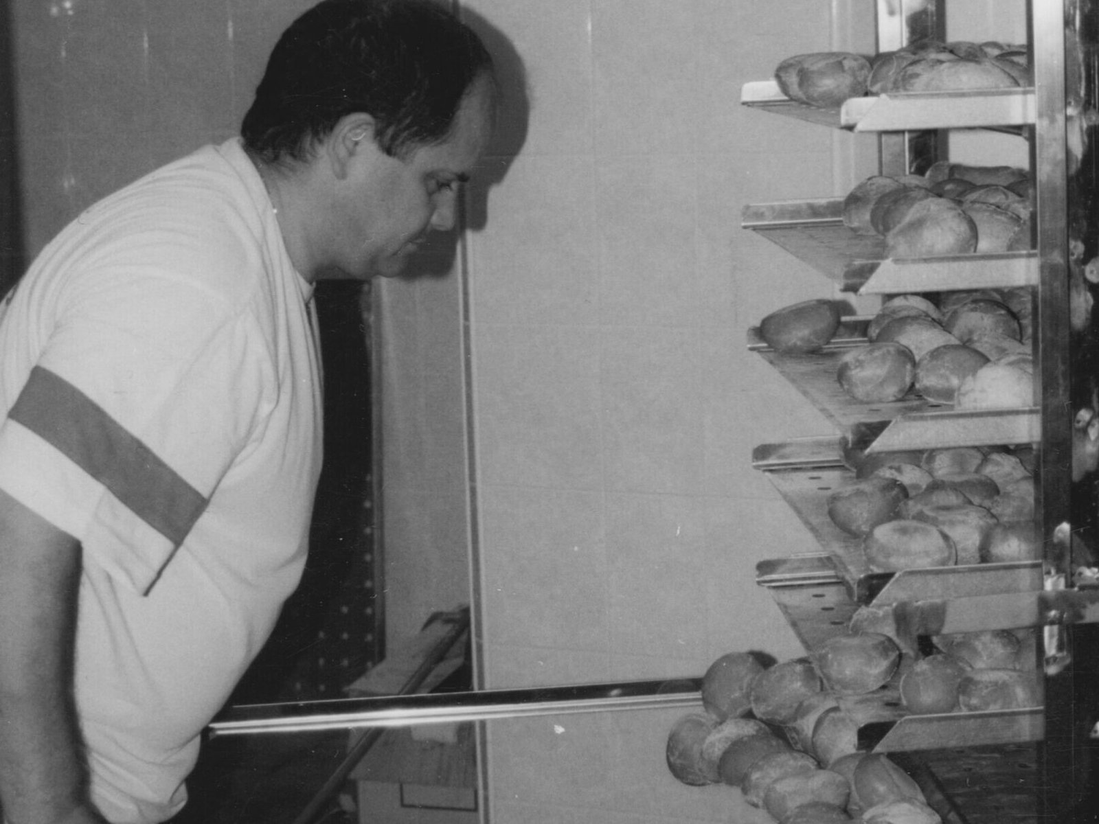 Maffia 1957 In un panificio di Como, un uomo in camicia a maniche corte usa un attrezzo a manico lungo per togliere i panini freschi dalle rastrelliere di metallo. Un cestino di plastica pieno di altri panini è appoggiato sul pavimento piastrellato lì vicino. Panetteria Pasticceria Caffetteria Como e Provincia