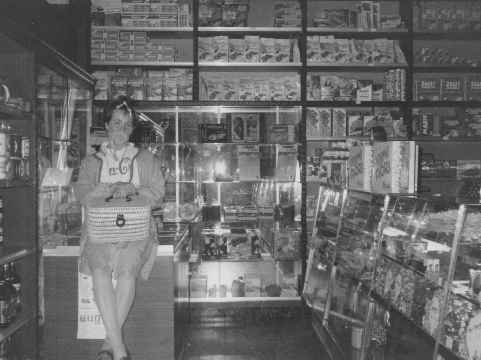 Maffia 1957 Una donna è in piedi e sorride all'interno di un piccolo negozio di alimentari, tenendo in mano un cestino. Gli scaffali dietro di lei sono pieni di vari prodotti alimentari confezionati, tra cui il pane artigianale Como, mentre le vetrine espongono altri articoli. La foto è in bianco e nero. Panetteria Pasticceria Caffetteria Como e Provincia