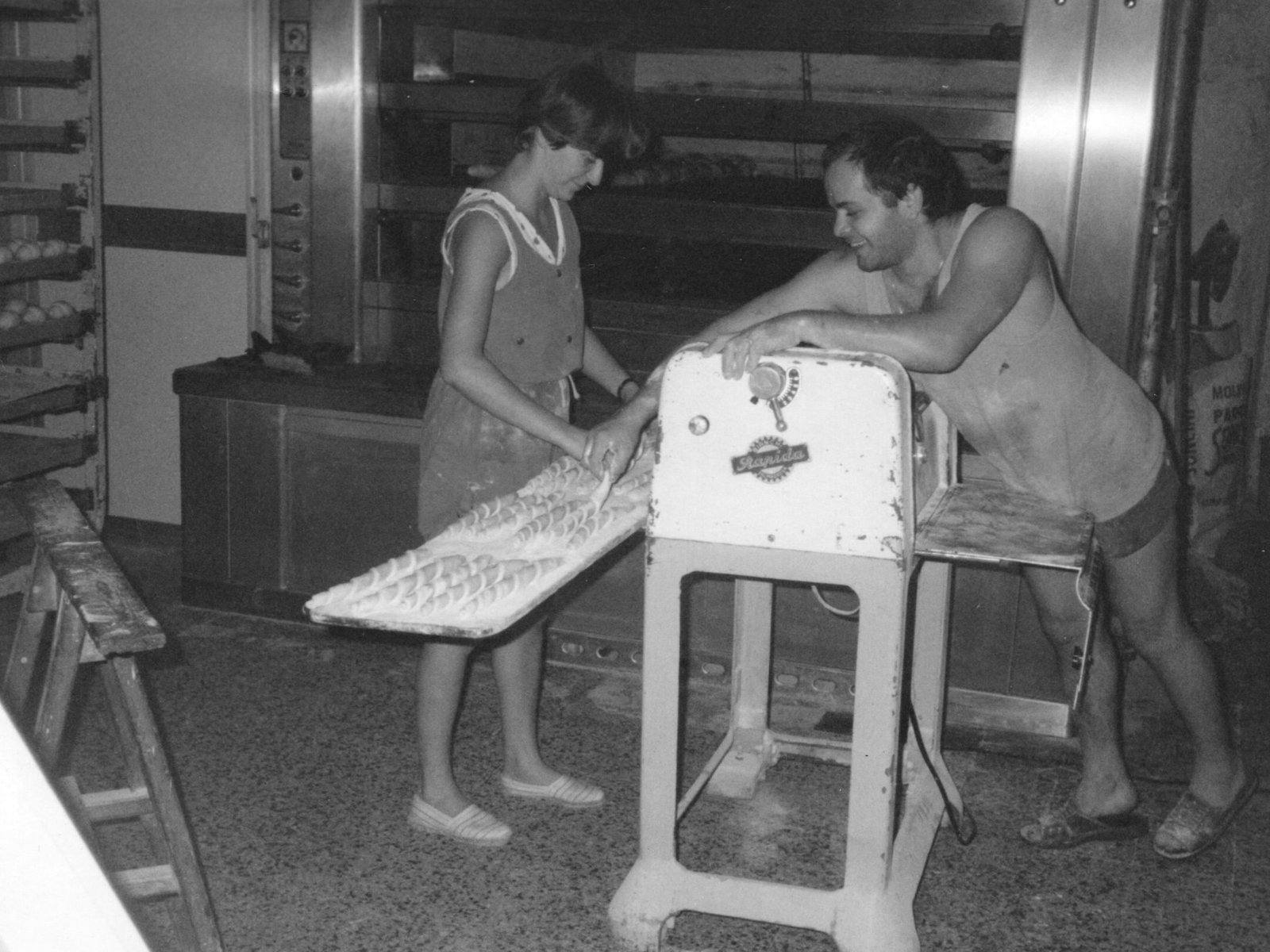 Maffia 1957 Due persone lavorano in una pasticceria di Como; una sistema l'impasto su un vassoio mentre l'altra si appoggia a una macchina. Sullo sfondo sono visibili forni industriali e scaffali pieni di teglie. Panetteria Pasticceria Caffetteria Como e Provincia